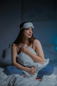 A serene photo of a woman wearing a sleep mask and holding a pillow, seated comfortably in a cozy bedroom.