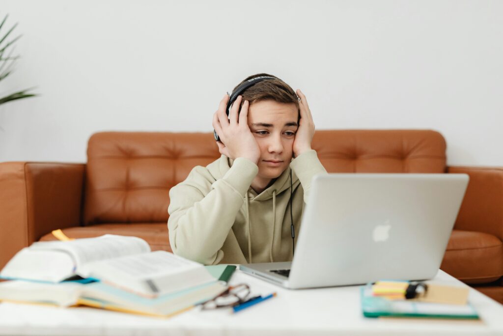 A young student wearing headphones, looking tired while studying with a laptop and books.